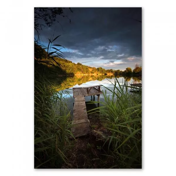 Holzsteg durch Schilfgras führt zu Plattform auf ruhigem See, Himmel strahlt in warmen Farben des Sonnenuntergangs. Holzsteg durch Schilfgras führt zu Plattform auf ruhigem See, Himmel strahlt in warmen Farben des Sonnenuntergangs.